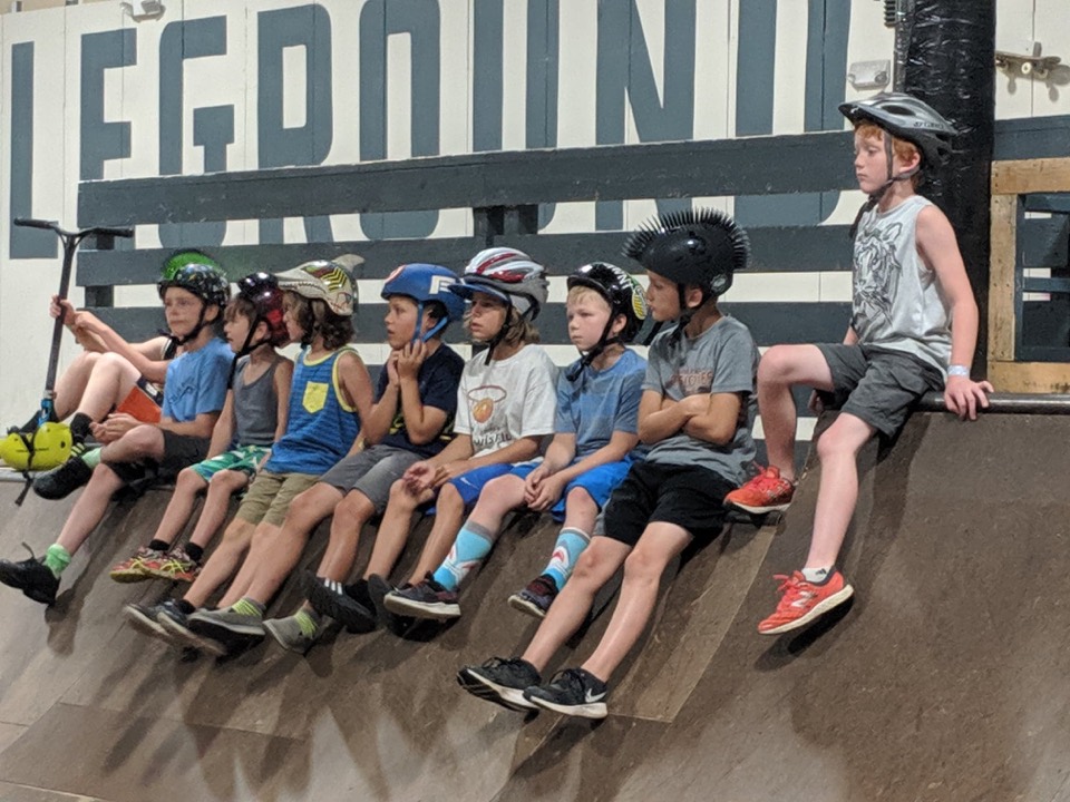 A row of kids in helmets sitting along the deck of a ramp, waiting for their turn