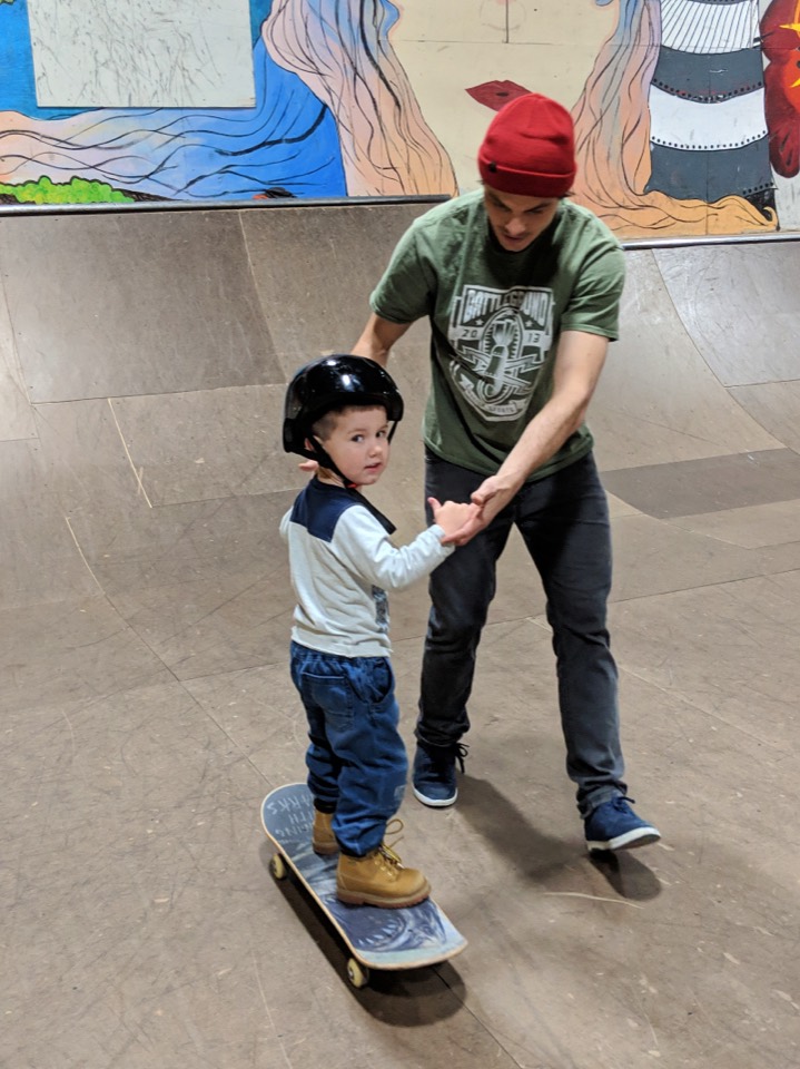 An adult crouching beside a small child in a helmet learning to balance on a board